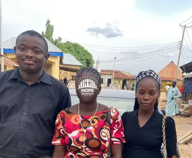 A woman, her face covered by the Hope Behind Bars Africa logo, stands on a street between a man and a woman.